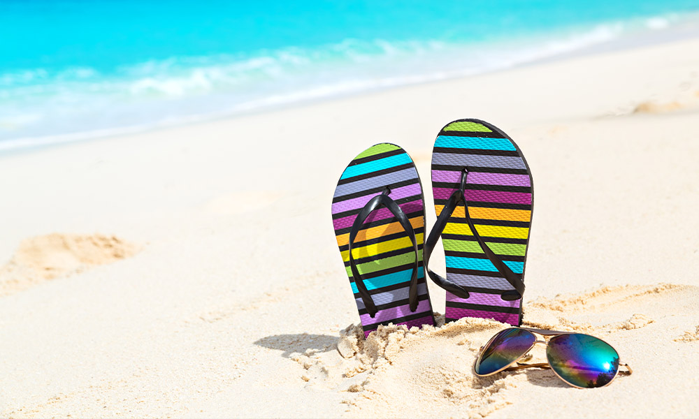 Q1 - State of the Market - Spring Break Flip Flop Sandals Sticking out of the sand on the beach with Sunglasses layed in front and the Ocean in the Background